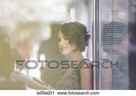 Stock Image - Young woman at bus stop reading newspaper and waiting for bus, New York, US. Fotosearch