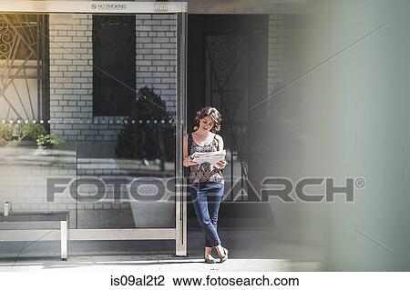 Stock Image - Young woman at bus stop reading newspaper and waiting for bus, New York, US. Fotosearch