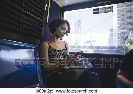 Stock Photography - Young woman commuting on bus listening to music on smartphone, New York, US. Fotosearch