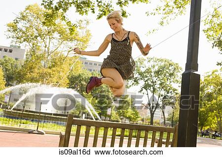 Stock Image - Young woman jumping mid air in park. Fotosearch