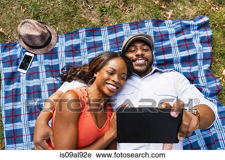 Stock Image - Couple lying on grass watching movie on tablet. Fotosearch