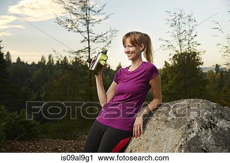 Stock Image - Female runner leaning against boulder in park drinking water. Fotosearch