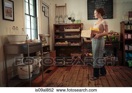 Stock Image - Female shop assistant carrying fruit and veg crate in country store. Fotosearch