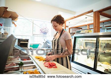 Stock Image - Female shop assistant slicing fruit in kitchen at country store. Fotosearch