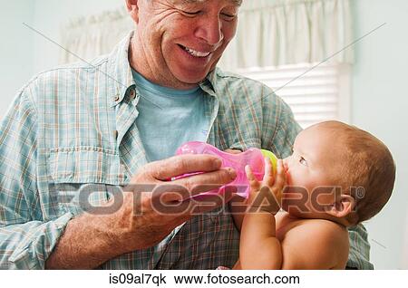 Stock Image - Grandfather bottle feeding baby granddaughter on lap in living room. Fotosearch