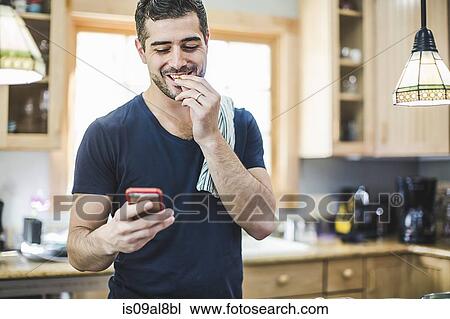 Stock Image - Man eating and using smartphone in kitchen. Fotosearch