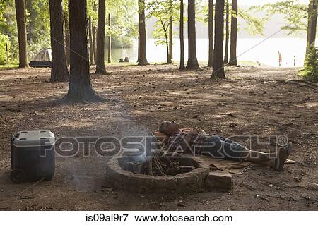 Stock Photo - Man lying down next to campfire in forest, Arkansas, USA. Fotosearch