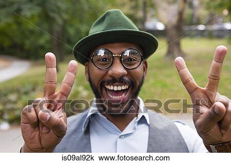 Stock Image - Man making peace sign smiling widely in park. Fotosearch