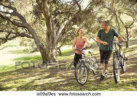 Stock Photograph - Mature couple chatting and pushing bicycles through park. Fotosearch