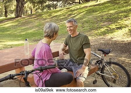 Stock Image - Mature cycling couple taking a break on park picnic table. Fotosearch