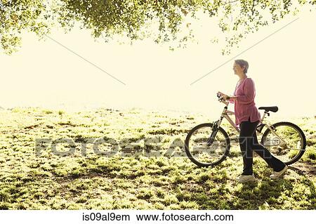 Picture - Mature woman pushing bicycle in park. Fotosearch