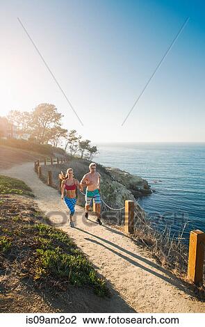 Stock Image - Mid adult man and young woman jogging on path by sea. Fotosearch