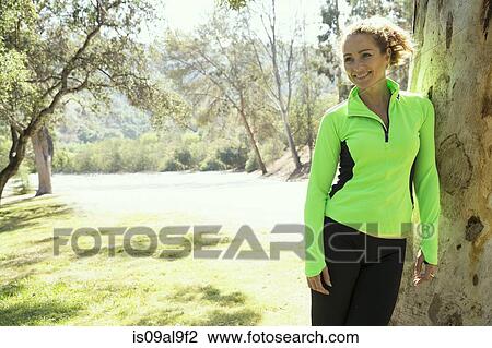 Stock Image - Portrait of mature female runner leaning against tree trunk in park. Fotosearch