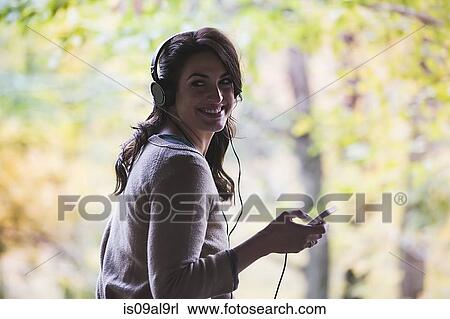 Stock Image - Portrait of young woman in forest listening to headphones. Fotosearch