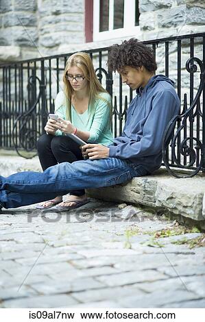 Two students sitting by railings using cell phone and tablet View Large Photo Image Stock Photo - Two students sitting by railings using cell phone and tablet. Fotosearch