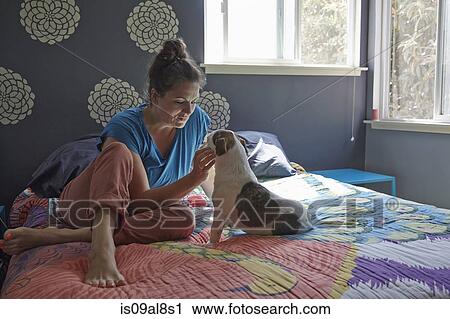 Stock Image - Woman sitting on bed petting dog. Fotosearch