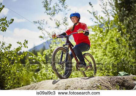 Stock Image - Young female bmx biker poised on edge of rock in forest. Fotosearch