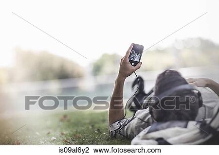 Stock Image - Young man lying on grass in park selecting music on smartphone. Fotosearch