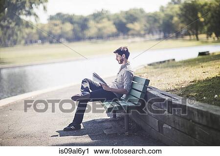 Stock Image - Young man reading newspaper on bench in park. Fotosearch
