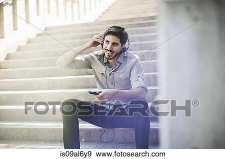 Stock Photo - Young man sitting on footbridge stairs listening to music on smartphone. Fotosearch
