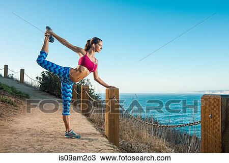 Stock Image - Young woman doing stretches on path by sea. Fotosearch