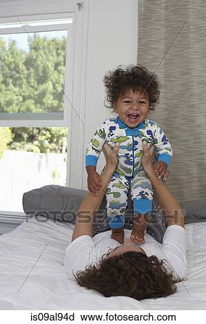 Stock Image - Young woman lying on bed holding up toddler son. Fotosearch