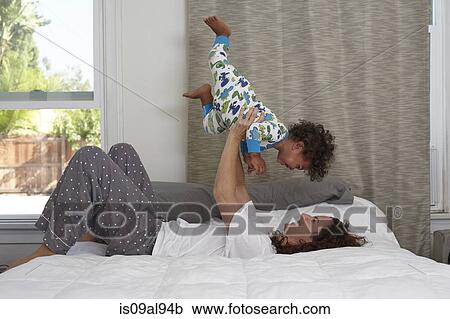 Stock Image - Young woman lying on bed lifting up toddler son. Fotosearch