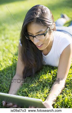Stock Image - Young woman lying on grass using digital tablet. Fotosearch