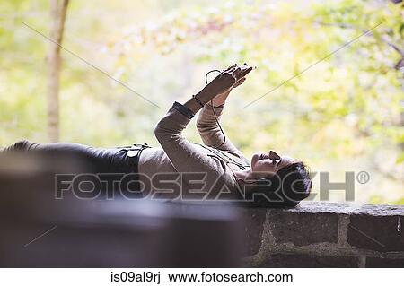 Stock Image - Young woman lying on wall holding up smartphone whilst listening to headphones. Fotosearch