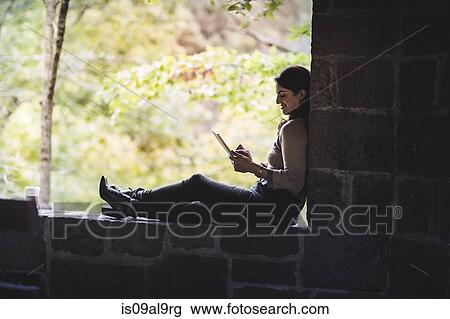 Stock Image - Young woman sitting on wall in forest looking at digital tablet. Fotosearch