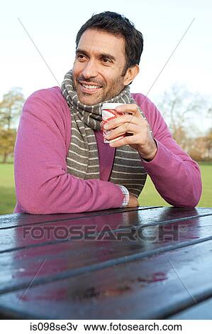 A man sitting at a park table holding a hot drink View Large Photo Image Stock Photo - A man sitting at a park table holding a hot drink. Fotosearch