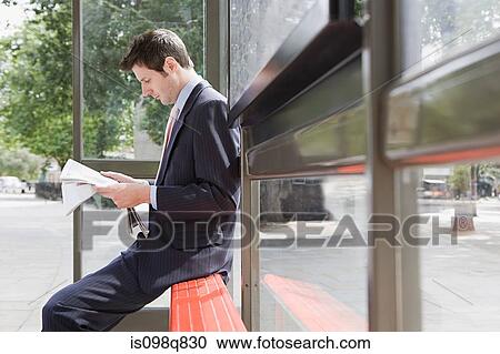 Stock Image - Businessman reading newspaper at bus stop. Fotosearch