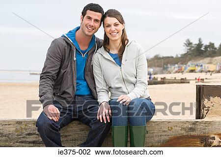 Stock Image - Couple sitting on a fence at the beach. Fotosearch