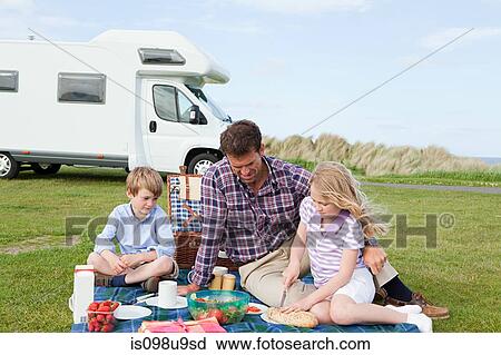 Stock Image - Father and children having picnic by caravan. Fotosearch