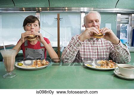 Girl and old man eating burgers in a diner View Large Photo Image Stock Photo - Girl and old man eating burgers in a diner. Fotosearch