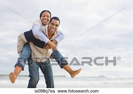 Stock Image - Man giving woman piggyback by the sea. Fotosearch