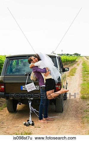 Stock Photograph - Newlywed couple kissing by vehicle. Fotosearch