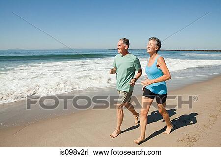Stock Image - Senior couple jogging on beach. Fotosearch