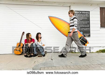 Picture - Two girls watching young man carrying surfboard. Fotosearch