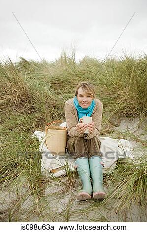 Stock Image - Woman sitting on dune with hot drink. Fotosearch