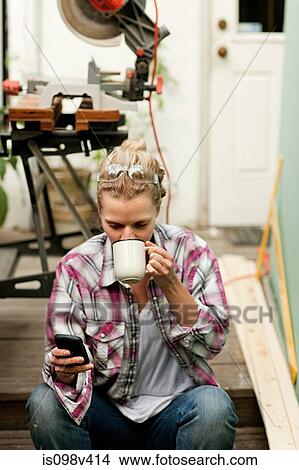 Picture - Woman sitting on steps with smartphone drinking hot drink. Fotosearch