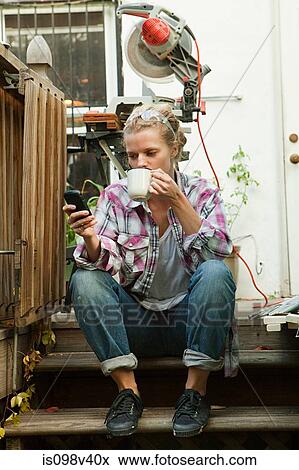 Stock Photo - Woman sitting on steps with smartphone drinking hot drink. Fotosearch