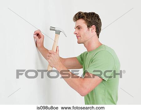 Young man hammering nail into wall View Large Photo Image Stock Image - Young man hammering nail into wall. Fotosearch