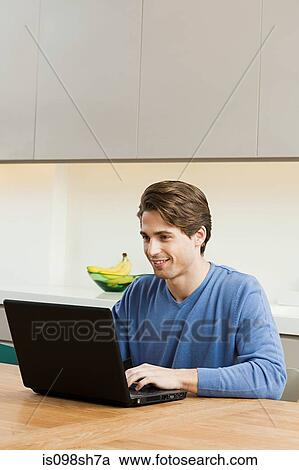 Young man sitting at table with laptop View Large Photo Image Stock Image - Young man sitting at table with laptop. Fotosearch