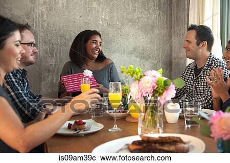 Stock Image - "Group of friends at dinner table, Young woman holding wrapped gift". Fotosearch