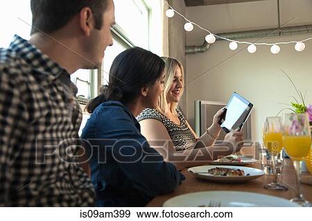 Stock Photo - "Group of friends at dinner table, looking at digital tablet screen". Fotosearch