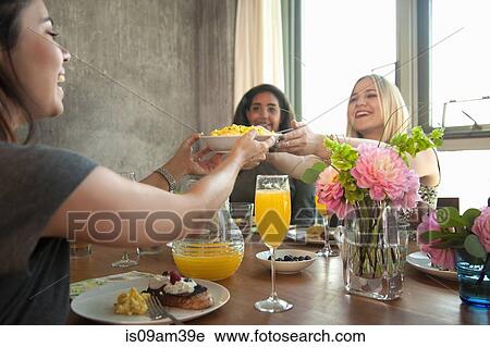 Stock Image - "Group of friends having meal at table, young woman passing plate to friend". Fotosearch