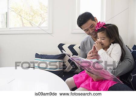 Stock Image - Mature man and daughter reading storybook on sofa. Fotosearch