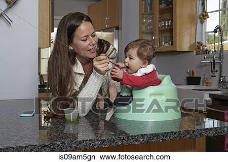 Picture - Mother feeding baby girl on kitchen counter. Fotosearch