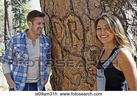 Stock Image - "Portrait of young couple in forest next to heart carving on tree trunk, Los Angeles, California, USA". Fotosearch